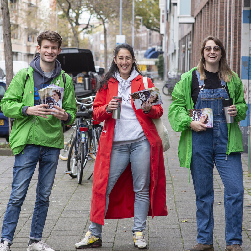 Drie vrijwilligers kijken lachend in de camera tijdens een flyeractie